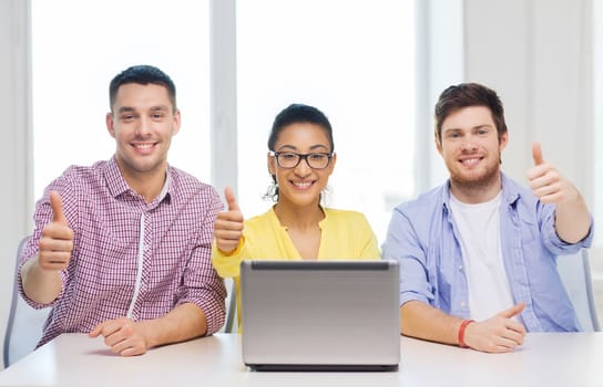 education, technology, business, startup and office concept - three smiling colleagues with laptop in office showing thumbs up