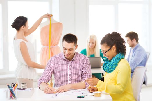 startup, education, fashion and office concept - smiling designers drawing sketches and adjusting dress on mannequin in office