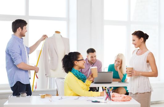 startup, education, fashion and office concept - smiling female designers having a coffee break in office with team on the back working