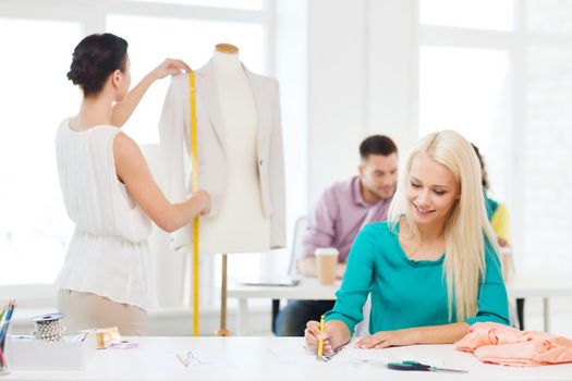 startup, education, fashion and office concept - smiling designers drawing sketches and measuring jacket on mannequin in office