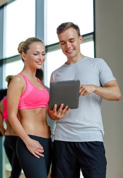 fitness, sport, exercising, technology and diet concept - smiling young woman and personal trainer with tablet pc computer in gym