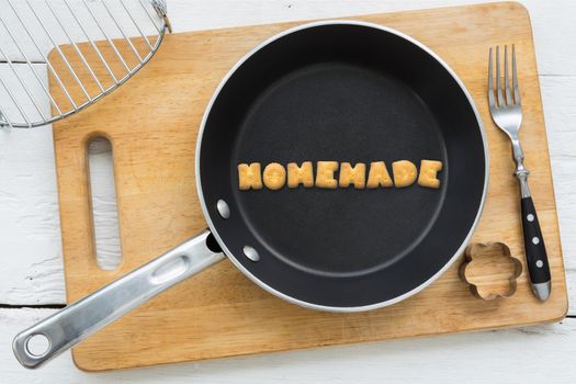 Top view of letter collage made of biscuits. Word HOMEMADE putting in black frying pan. Other cooking equipments: fork, cookie cutter and chopping board putting on white wooden table, vintage style image.