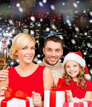 christmas, holidays, family and people concept - happy mother, father and little girl in santa helper hat with gift boxes over snowy night city background