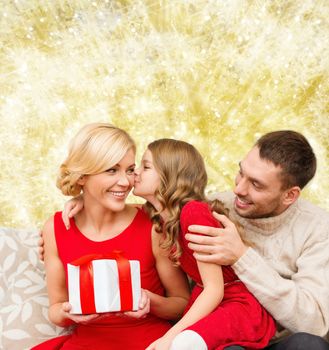 christmas, holidays, family and people concept - happy mother, father and little girl with gift box kissing over yellow lights background