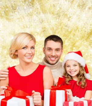 christmas, holidays, family and people concept - happy mother, father and little girl in santa helper hat with gift boxes over yellow lights background