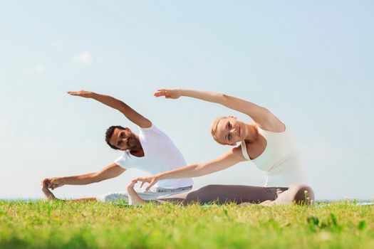 fitness, sport, friendship and lifestyle concept - smiling couple making yoga exercises sitting on mats outdoors