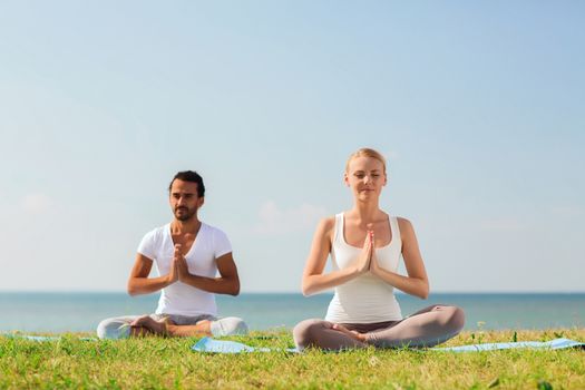 fitness, sport, friendship and lifestyle concept - smiling couple making yoga exercises sitting on mats outdoors