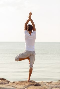 fitness, sport, people and lifestyle concept - young man making yoga exercises on beach from back