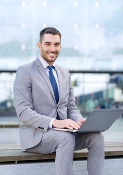 business, education, technology and people concept - smiling businessman working with laptop computer on city street