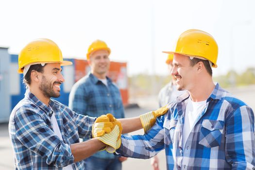 business, building, teamwork, gesture and people concept - group of smiling builders in hardhats greeting each other with handshake outdoors