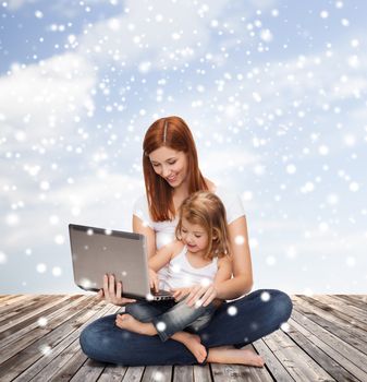 childhood, parenting, people and technology concept - happy mother with little girl with laptop computer over wooden floor and blue sky background
