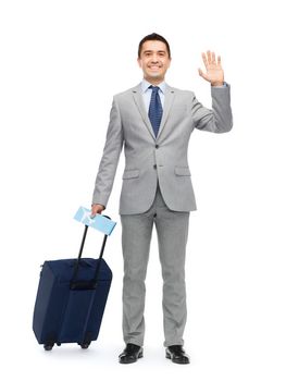 business trip, traveling, luggage and people concept - happy businessman in suit with travel bag and air ticket waving hand