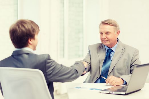 business, technology and office concept - older man and young man shaking hands in office