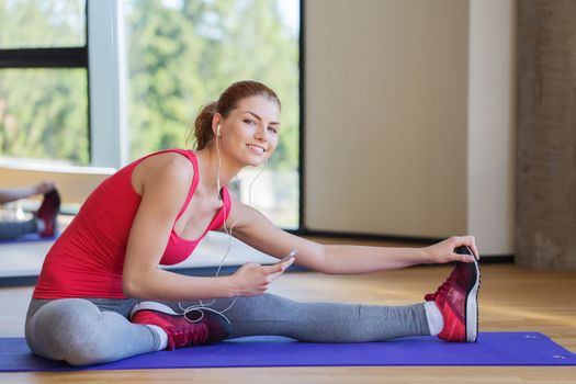 fitness, sport, people, technology and lifestyle concept - smiling woman with smartphone and earphones stretching in gym