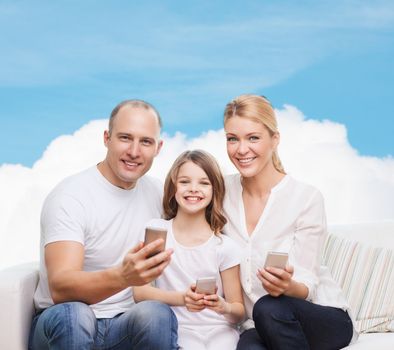 family, technology and people concept - smiling mother, father and little girl with smartphones over blue sky and white cloud background