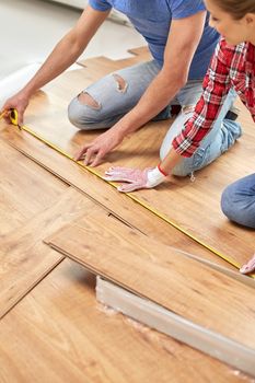 repair, building, flooring and people concept - happy couple with ruler measuring parquet board at home