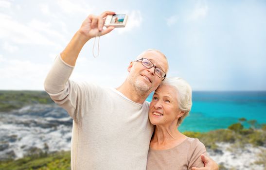 age, tourism, travel, technology and people concept - senior couple with camera taking selfie on street over beach background