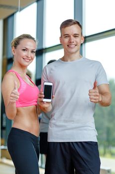 fitness, sport, advertising, technology and diet concept - smiling young woman and personal trainer with smartphone in gym showing thumbs up