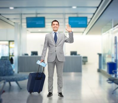 business trip, traveling, luggage and people concept - happy businessman in suit with travel bag and air ticket waving hand over airport background
