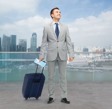 business trip, traveling, luggage and people concept - happy businessman in suit with travel bag and air ticket over city background