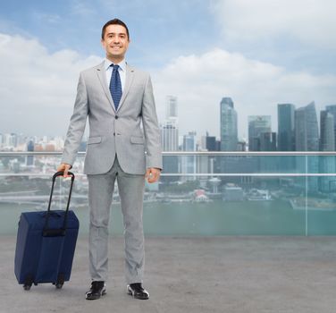 business trip, traveling, luggage and people concept - happy businessman in suit with travel bag over city background