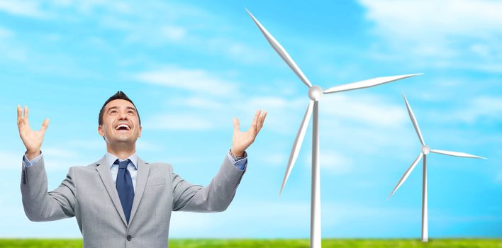 business, people, alternative energy and development concept - happy businessman in suit with raised hands laughing and looking up over blue sky and windmills background