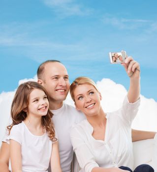 family, technology and people - smiling mother, father and little girl making selfie with camera over blue sky and white cloud background