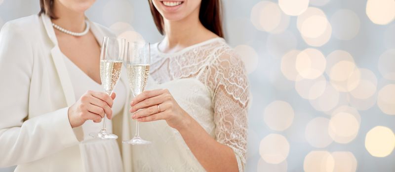 people, homosexuality, same-sex marriage, celebration and love concept - close up of happy married lesbian couple holding and clinking champagne glasses over holidays lights background