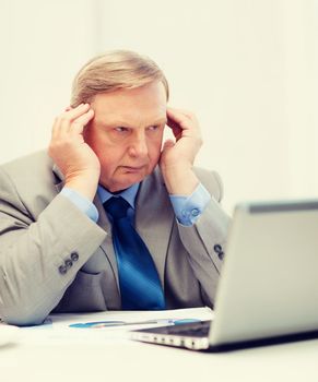 business, technology and office concept - upset older businessman with laptop and charts in office