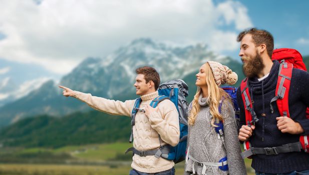 adventure, travel, tourism, hike and people concept - group of smiling friends with backpacks pointing finger over alpine mountains background