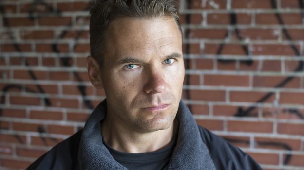 Closeup portrait of Caucasian man with green hazel eyes staring into camera with serious expression on blurred background of red brick wall with grafitti