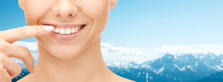 dental health, beauty, hygiene and people concept - close up of smiling woman face pointing to teeth over blue mountains background