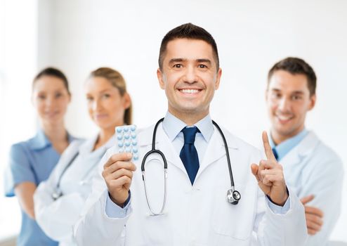 healthcare, profession, people and medicine concept - smiling male doctor in white coat with tablets pointing his finger up over group of medics at hospital background