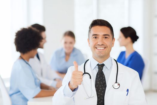 clinic, profession, people and medicine concept - happy male doctor over group of medics meeting at hospital showing thumbs up gesture