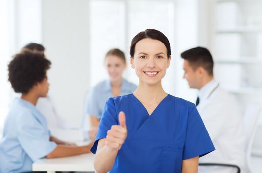 clinic, profession, people and medicine concept - happy female doctor or nurse over group of medics meeting at hospital showing thumbs up gesture