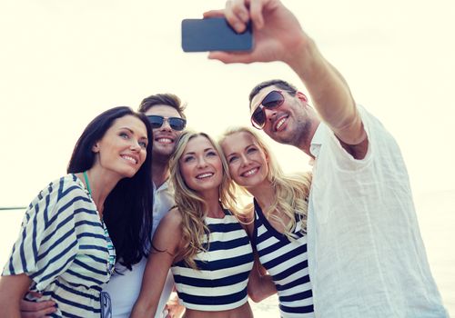 summer, sea, tourism, technology and people concept - group of smiling friends with smartphone on beach photographing and taking selfie