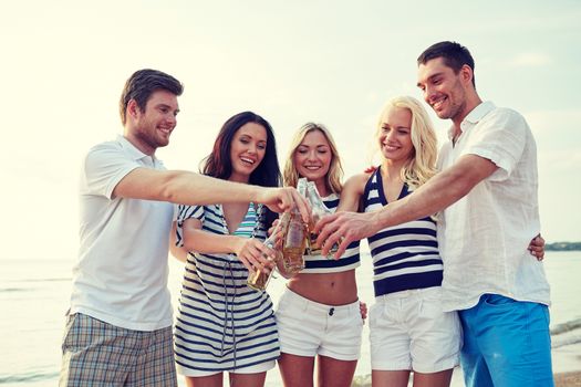 summer, holidays, tourism, drinks and people concept - group of smiling friends clinking bottles of beer or cider on beach