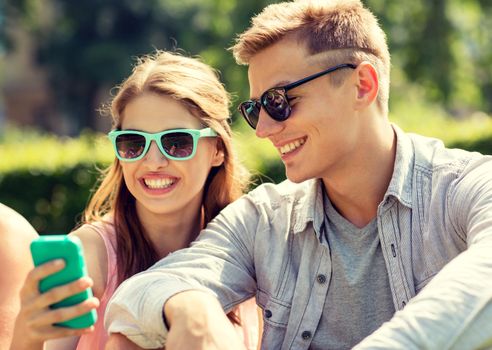 friendship, leisure, summer, technology and people concept - group of smiling friends with smartphone sitting on grass in park