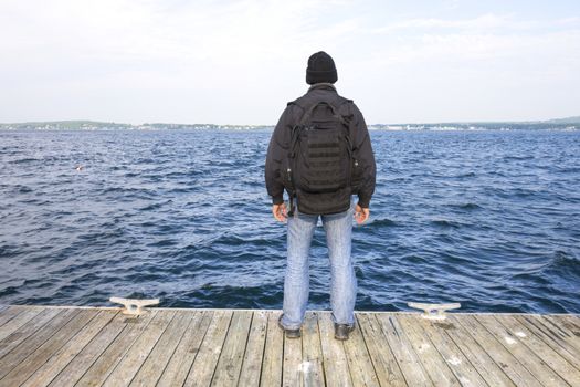 Back side of man with black backpack and jacket standing at end of wooden dock overlooking choppy waters of bay in Rockland, Maine