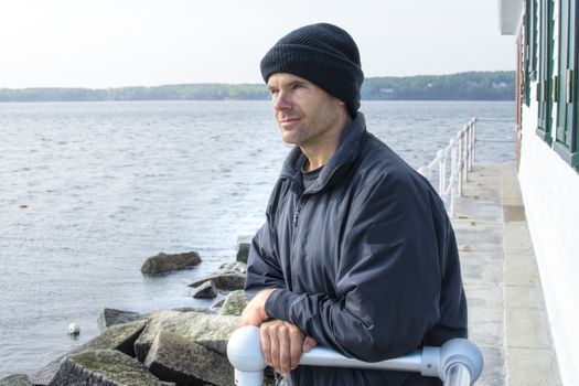 Handsome Caucasian man dressed in jacket and black cap leans against railing of lighthouse at end of breakwater in Rockland, Maine as he gazes peacefully into the distant ocean