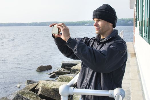 Caucasian man dressed in black jacket and beanie takes cell phone photo of scenic view of bay at the lighthouse at end of breakwater in Rockland, Maine