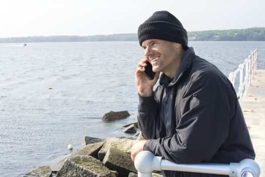 Handsome Caucasian man smiles while talking on cell phone as he leans against railing at end of breakwater of bay in Rockland, Maine