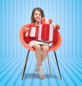 people, christmas, holidays, presents and childhood concept - happy little girl with gift boxes sitting on chair over blue striped background