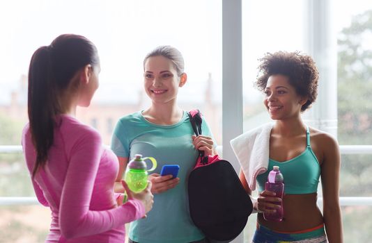 fitness, sport, training and lifestyle concept - group of happy women with bottles of water, smartphone and bag talking in gym