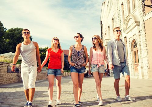 friendship, leisure, summer, gesturer and people concept - group of smiling friends walking and holding hands in city