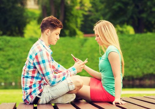 summer, vacation, technology, addiction and friendship concept - couple with smartphones sitting on bench in park
