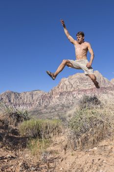 Muscular shirtless Caucasian man wearing shorts and sandals runs and jumps high in air over bushes in harsh desert landscape under clear sunny blue sky in Red Rock Canyon, Las Vegas