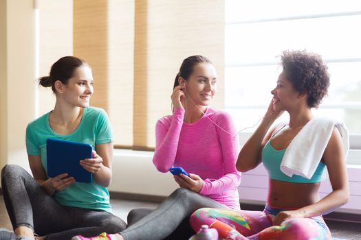 fitness, sport, technology and lifestyle concept - group of happy women with smartphone, earphones and tablet pc computer listening to music in gym