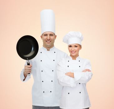 cooking, profession, teamwork, kitchenware and people concept - happy chefs or cooks couple with frying pan over beige background
