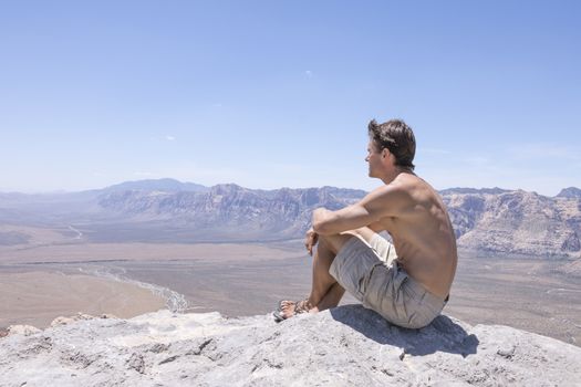 Wide angle of shirtless muscular Caucasian man in shorts and sandals sitting on rocky summit of mountain overlooking expanse of desert under sunny clear sky in Red Rock Canyon, Nevada